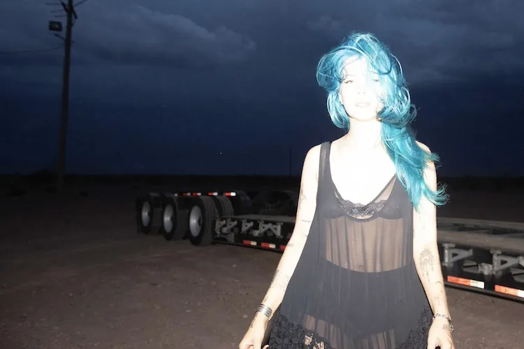 A figure with bright blue hair in a sheer black slip standing on a dark roadside at night, a truck trailer behind her