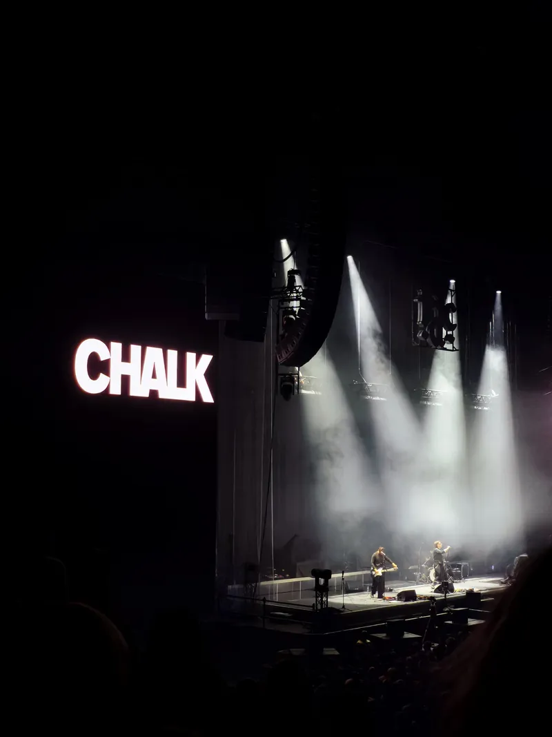 Chalk on a vast dark stage, two small figures under white spotlights cutting through the haze, with the name CHALK glowing in white capitals on the screen to the left