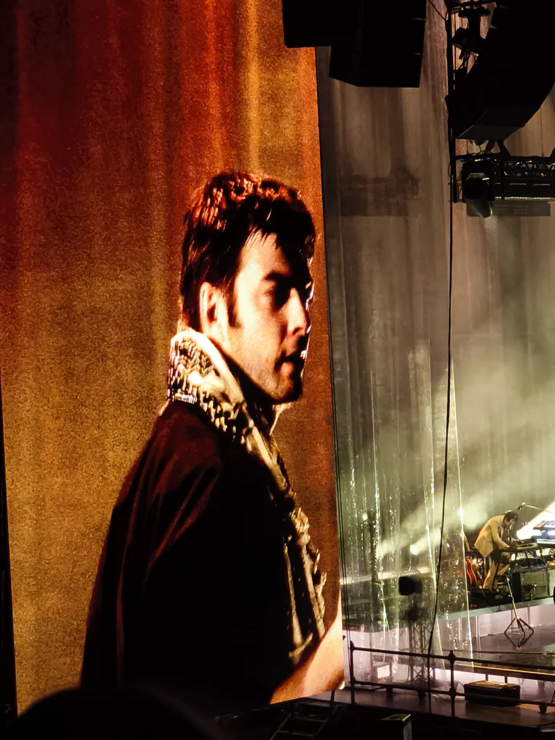 On the giant screen, a band member seen in profile with a keffiyeh around his neck, lit amber against the shimmering curtain, the keyboard player small on the stage behind