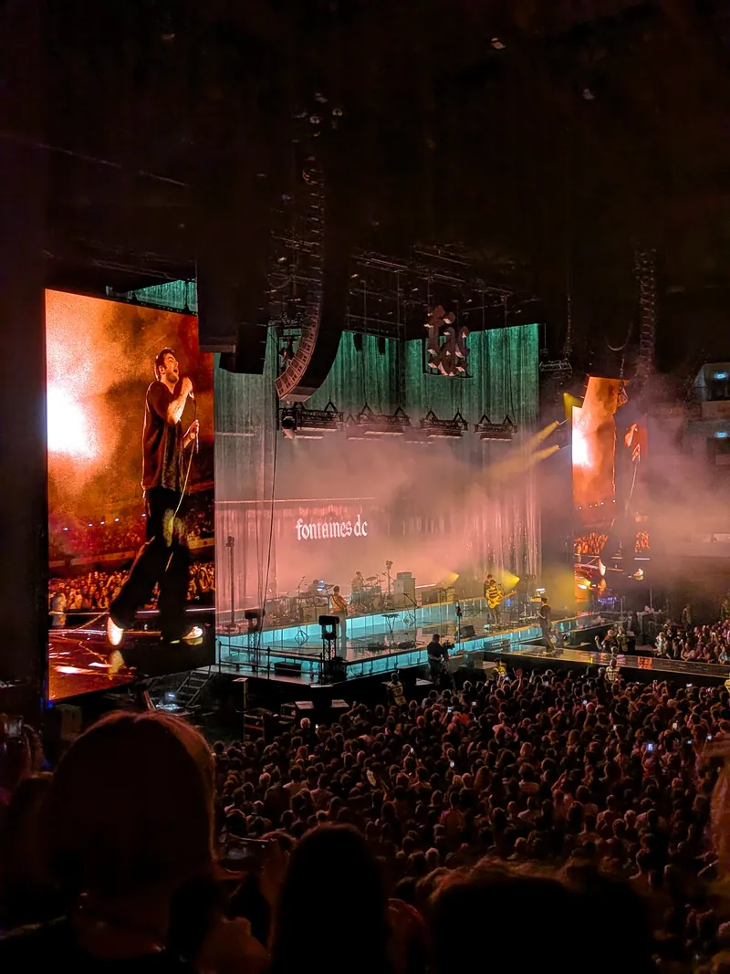The stage seen from the stands: the singer on the giant screen glowing orange on the left, the band small under fontaines dc projected on the green curtain, the gothic f.d.c sign hanging above, and a packed floor in the foreground