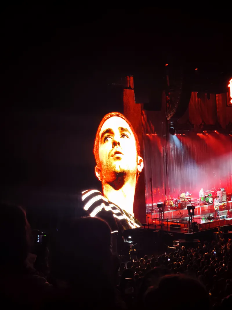 Carlos O’Connell on the giant screen, face lit orange and looking up, in a black and white striped top, the stage bathed in red light beside him