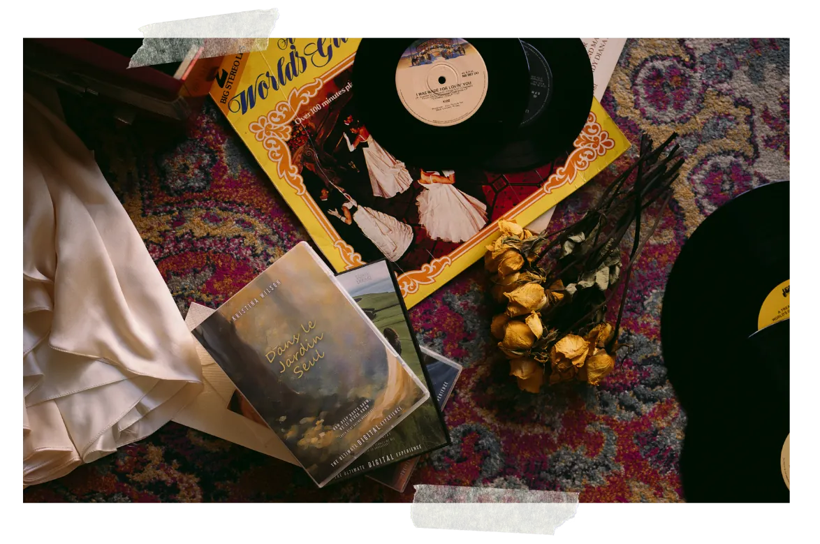 A flat-lay on a patterned rug — vinyl singles out of their sleeves, a cassette, dried yellow roses and cream satin