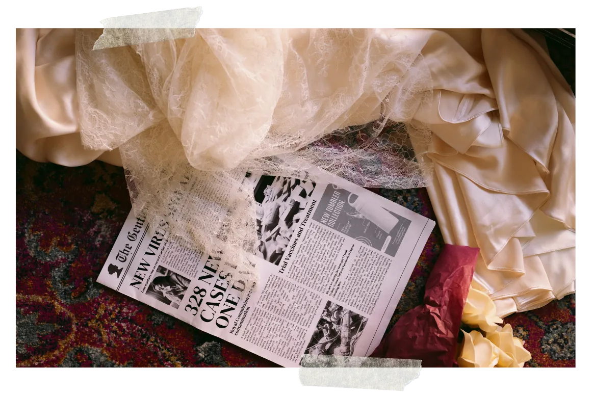 A flat-lay — cream silk and lace spread over a patterned rug, an open newspaper page about an outbreak, a dried rose beside it