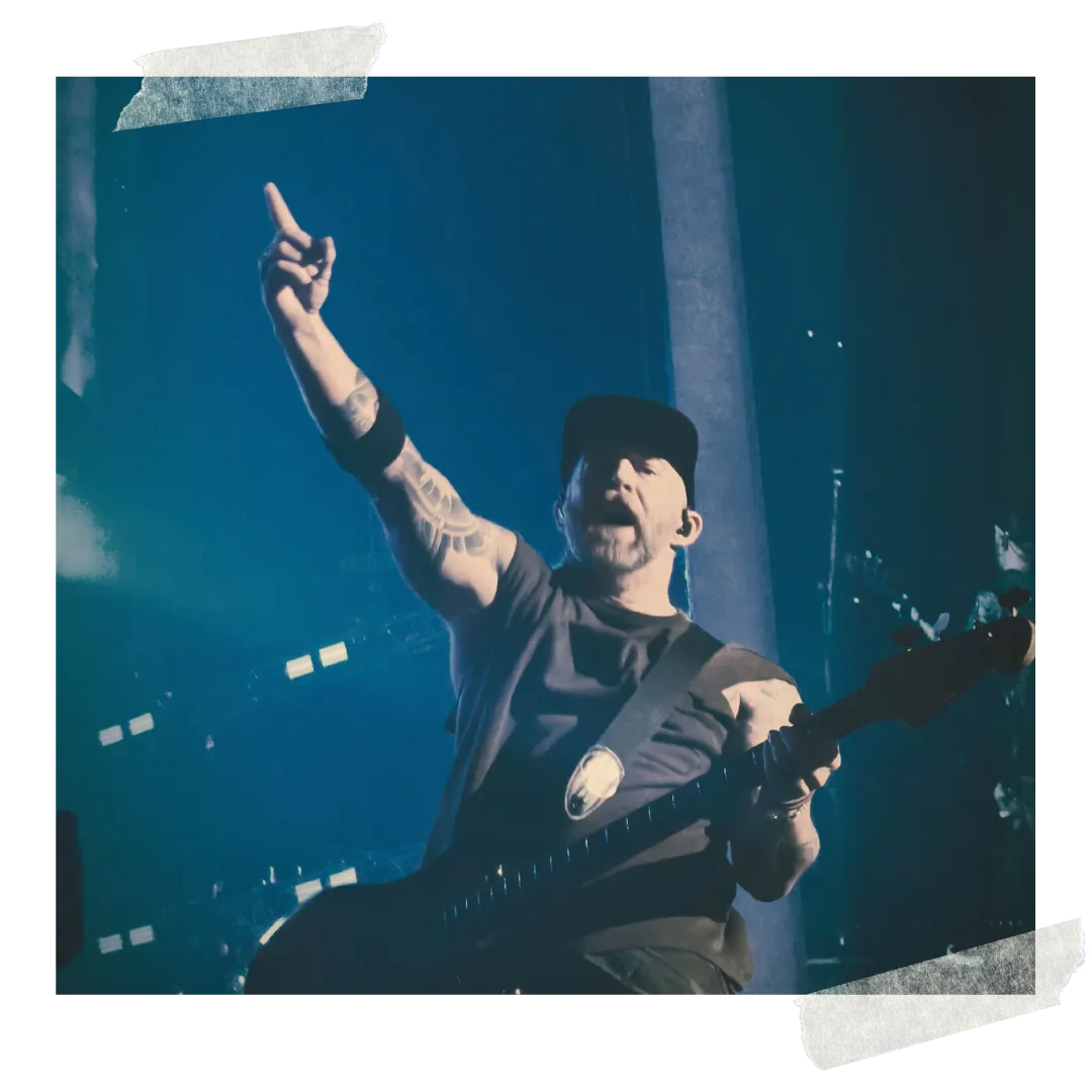 A guitarist pointing out to the crowd, his guitar slung low, under blue light