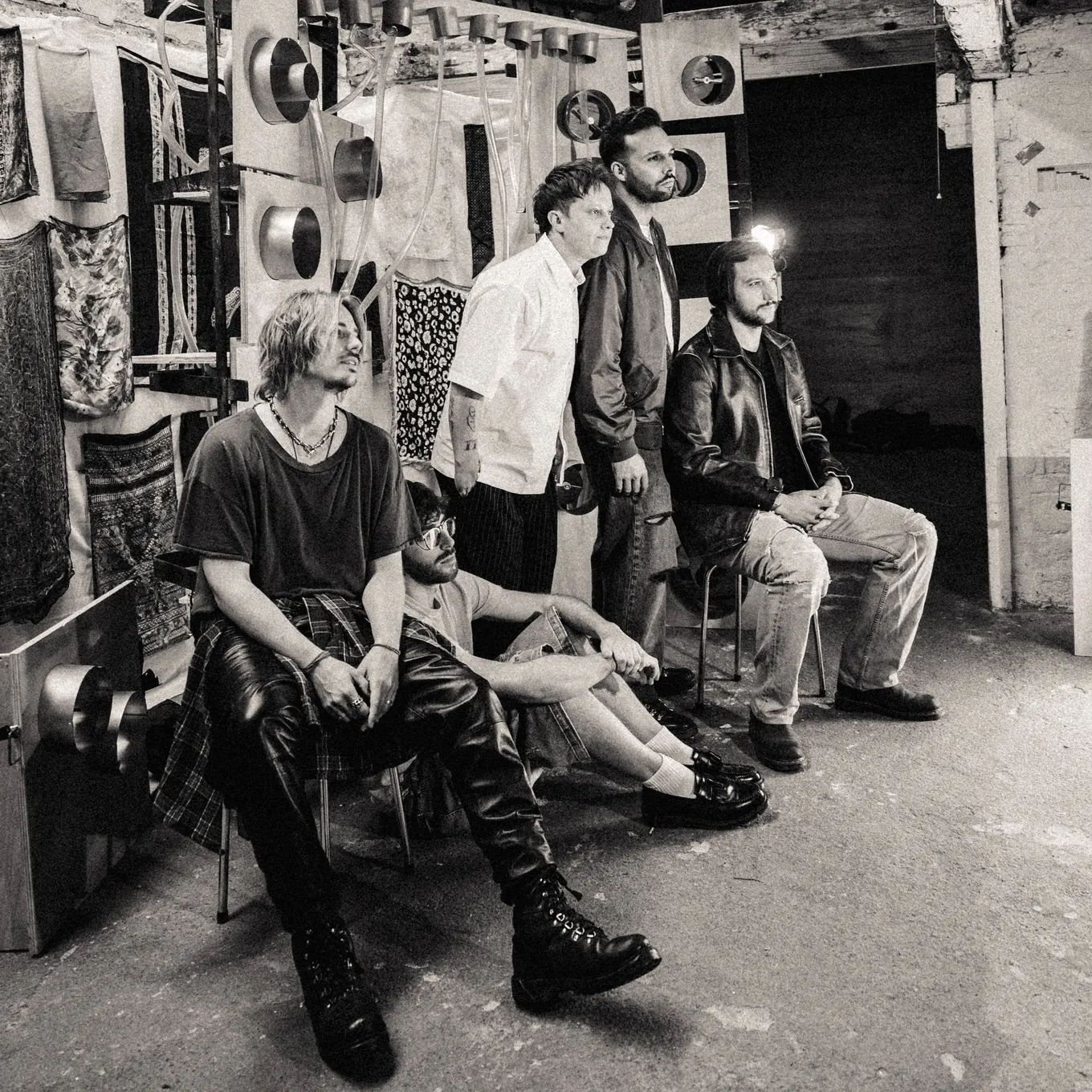 The five band members sitting and standing against a basement wall hung with fabric swatches and metal discs, black and white