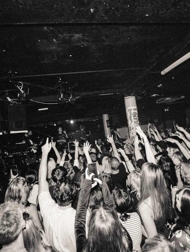 A packed club seen from the back of the room, hundreds of raised hands and phones facing a small lit stage, black and white
