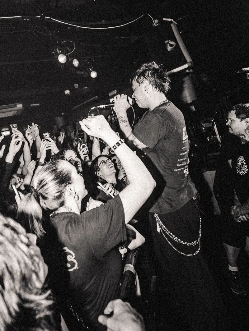 The singer standing on the barrier above the front row, microphone to his mouth, fans reaching up around him, black and white