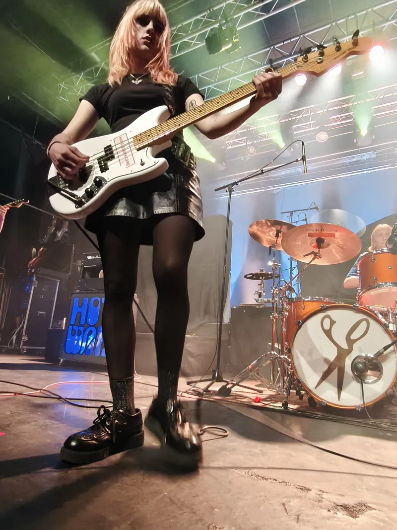 A bassist in a black top and short skirt, white bass raised, a drum kit with a pair of scissors painted on the kick drum behind