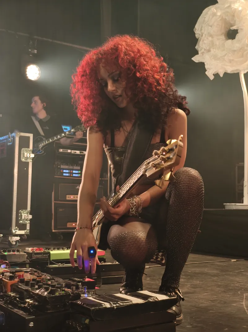 A bassist with a tall red afro crouching over her pedalboard, a gold bass across her knees