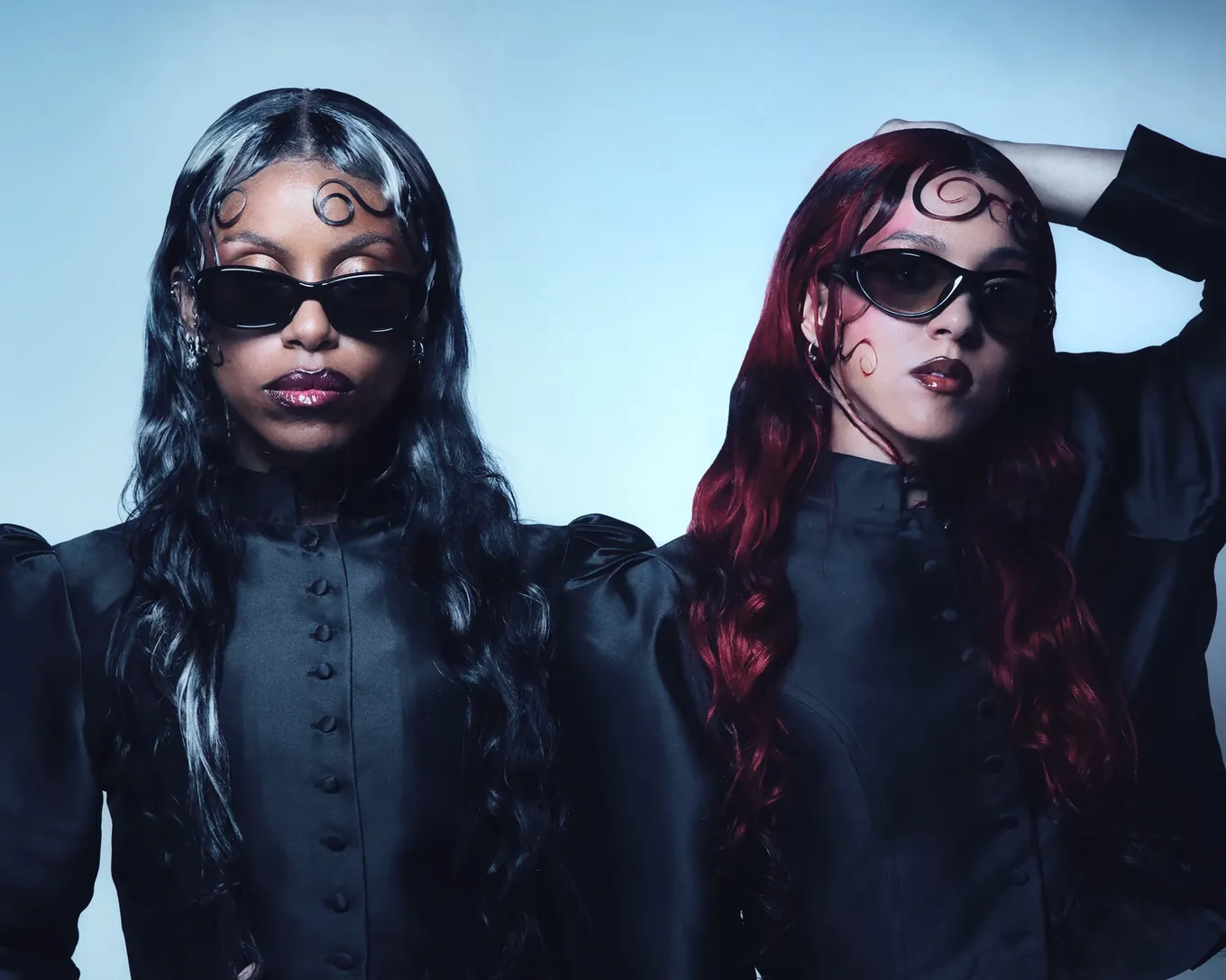 The duo against a pale blue sky in high-collared black, dark sunglasses, symbols drawn on their foreheads