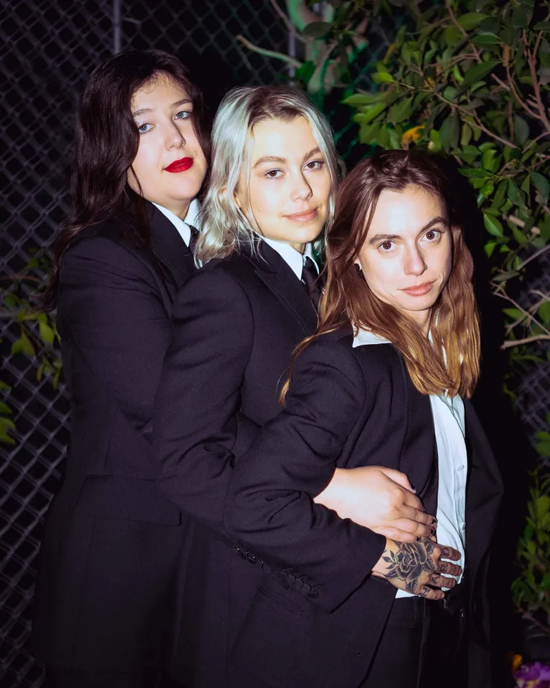 Three musicians in matching black suits standing close together in front of a chain-link fence