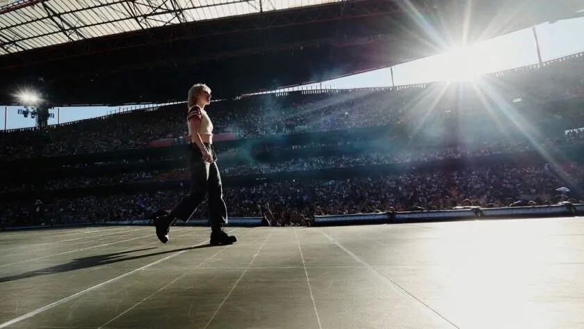 The singer walking a long stadium runway in low sun, the stands rising behind