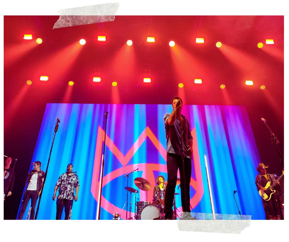 The band spread across a wide stage under red lights, the band's symbol on a blue and pink backdrop