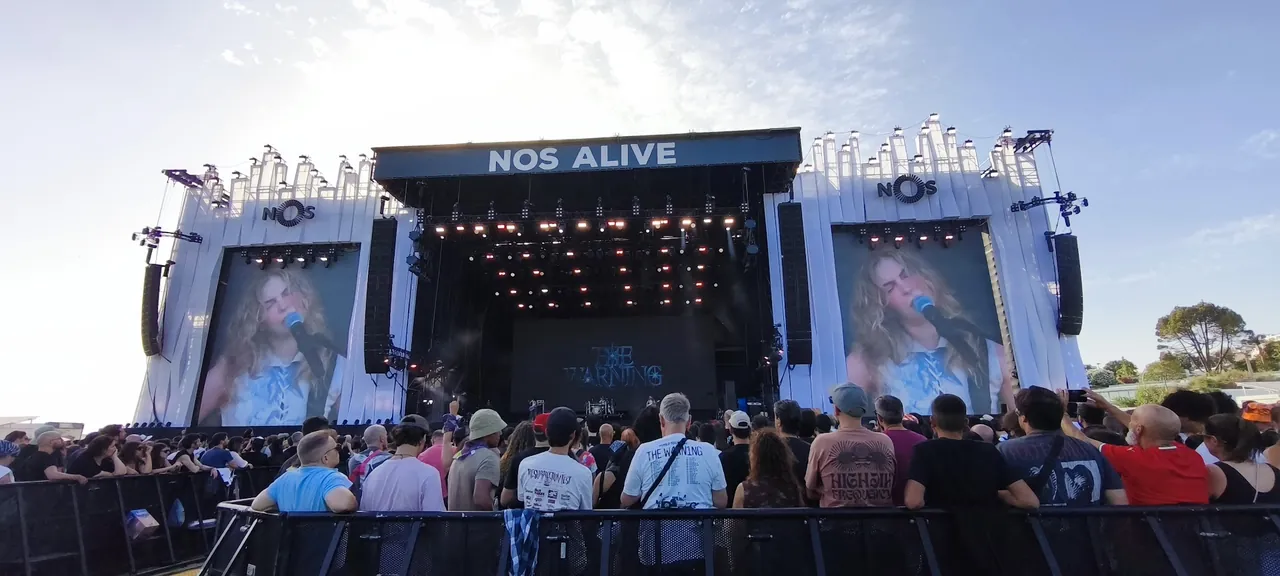 The NOS Alive main stage in late-afternoon light, THE WARNING on the backdrop and the singer on both side screens, a crowd pressed to the barrier below
