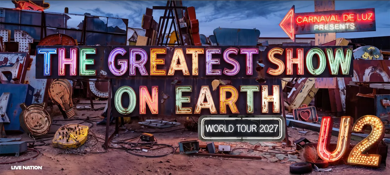 Tour poster for U2’s The Greatest Show On Earth, World Tour 2027: the title in huge rusted neon letters, each one a different colour, stands in a desert graveyard of old signs at dusk. A red neon arrow reads Carnaval De Luz presents; around it lie a giant lightbulb, a yellow lemon, a suitcase with a heart and a small Joshua tree sign, and a big red and gold U2 sign burns in the bottom corner