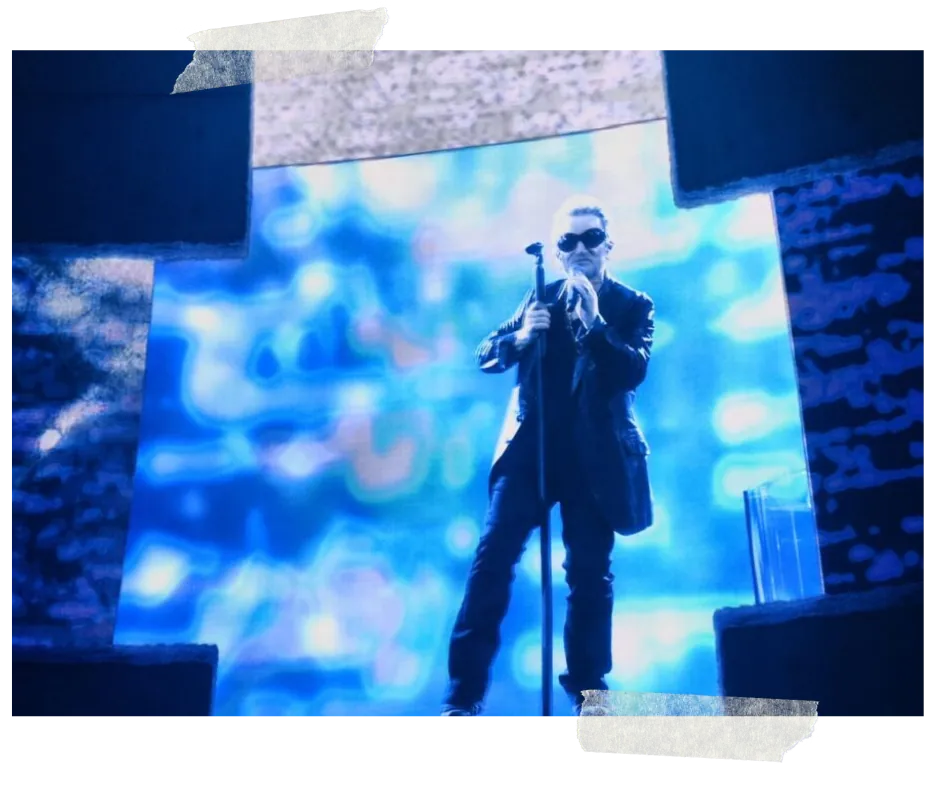 The singer in dark glasses on stage in front of a vast blue pixelated wall