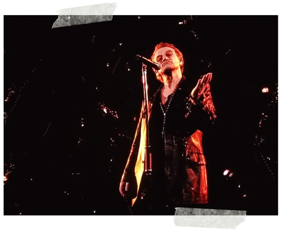 The singer in a long dark coat at the microphone, lit red against a black screen