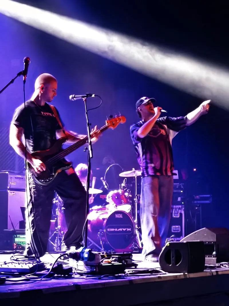 SNAYX on stage under purple light, the bassist mid-riff and the singer with one arm thrown out to the crowd