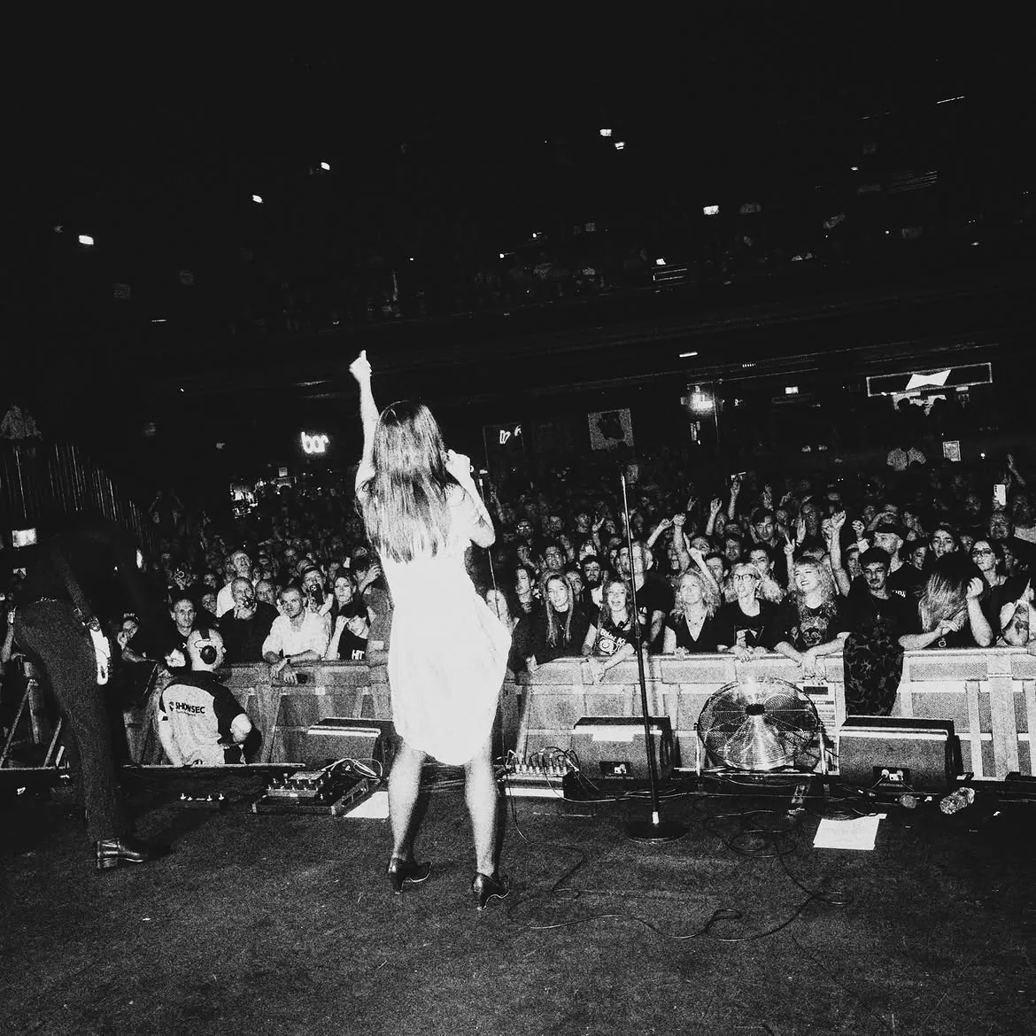 Seen from behind the singer at the front of the stage, one arm raised, facing a packed room with the balcony above, black and white