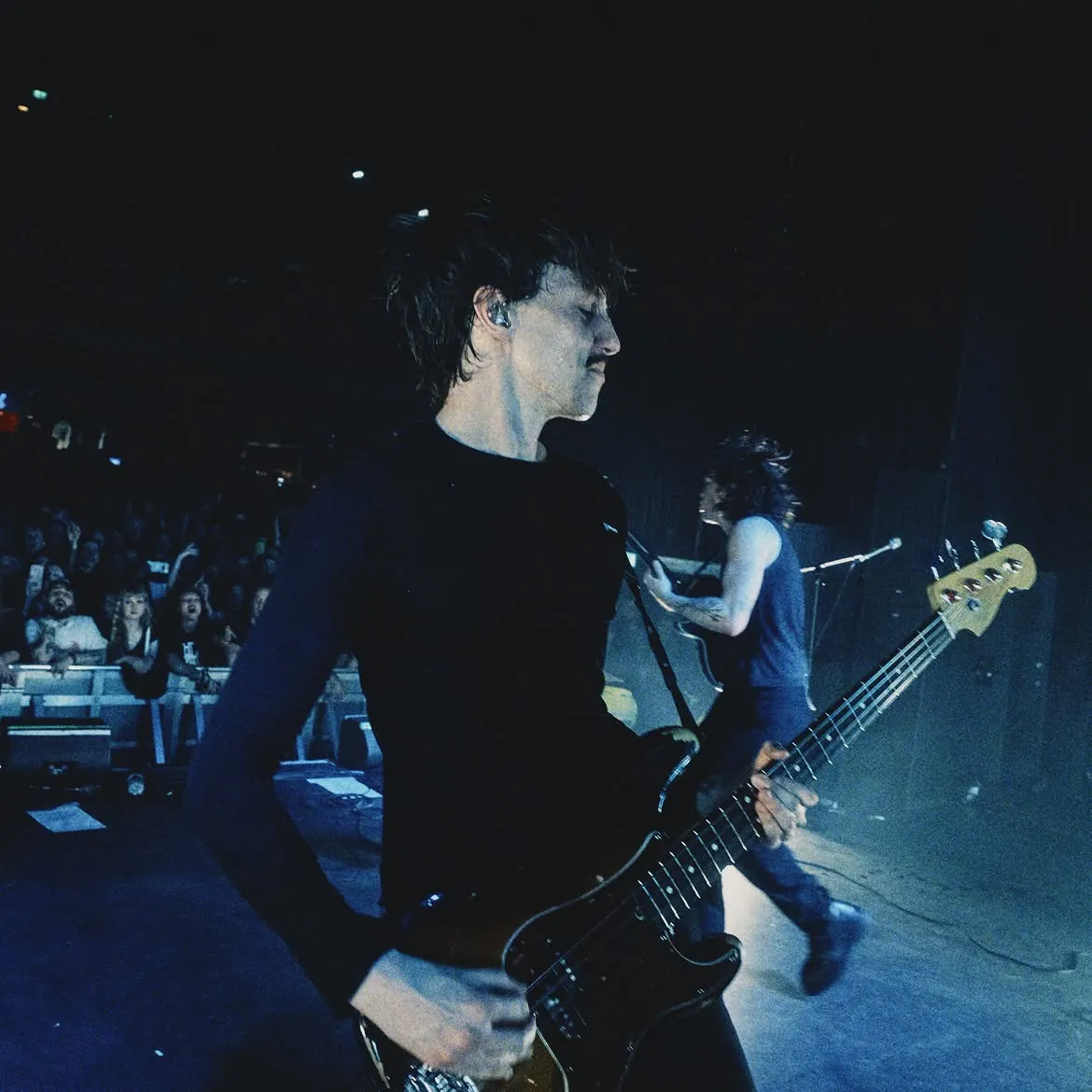 The guitarist in the foreground and the bassist walking behind him, the front row lit at the edge of frame, black and white