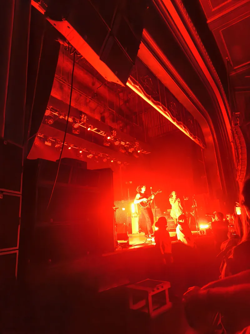 The acoustic set seen from the side of the room in deep red light, two figures on a small stage under the lighting rig
