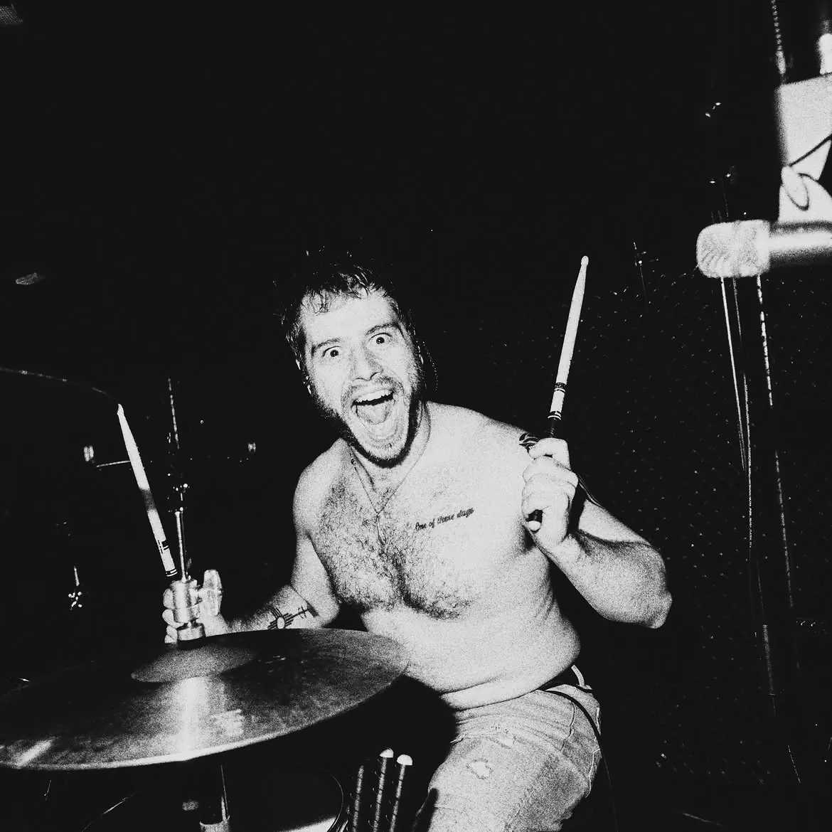 The drummer shirtless behind his kit, both sticks raised, grinning straight at the camera, black and white
