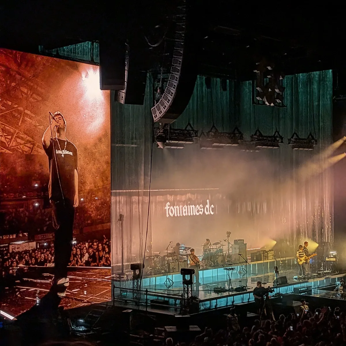Fontaines D.C. on stage at the MEO Arena: the singer standing on a giant screen bathed in orange light on the left, the band small on a lit stage in front of a shimmering green curtain with fontaines dc projected across it, the crowd in the foreground